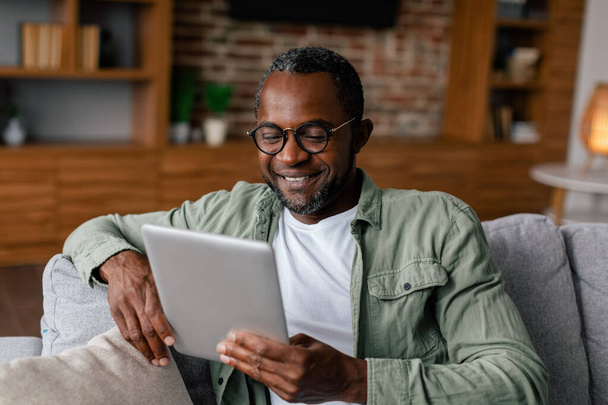 Fröhlicher erwachsener afrikanisch-amerikanischer Mann mit Brille und lässigem Videogucken auf dem Tablet, plaudernd im Wohnzimmer-Interieur. Neue App, Fernarbeit, Blog in sozialen Medien, neue Normalität während der Covid-19-Quarantäne - Foto, Bild