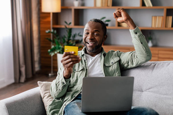 Spokojeně vzrušený dospělý africký Američan se raduje ze skvělého nákupu a vrácení peněz s notebookem a kreditní kartou v interiéru obývacího pokoje. Reklama a nabídka, on-line nakupování doma, vítězství a prodej - Fotografie, Obrázek