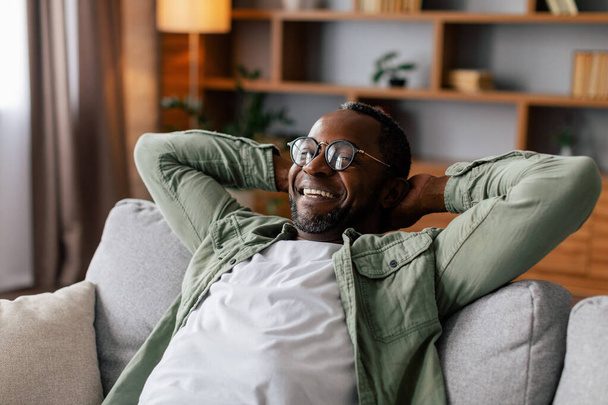 Homem afro-americano adulto satisfeito em óculos e descanso casual e relaxante, desfrutar de conforto, paz e tempo livre sozinho na sala de estar interior. Faça uma pausa em casa aconchegante durante a quarentena covid-19 - Foto, Imagem