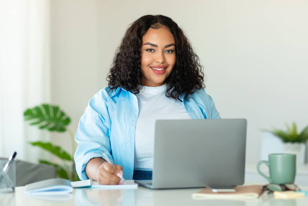 Щаслива афроамериканська бізнес леді використовує ноутбук приймаючи ноутбуки на робочому місці, посміхаючись камері позує сидячи в сучасному офісі. Успішна концепція бізнес-кар'єри. Вибірковий фокус
 - Фото, зображення