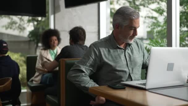 Homme d'âge moyen se préparant à travailler l'ajustement chemise et l'organisation manteau et articles en face de l'ordinateur portable assis au café - Séquence, vidéo