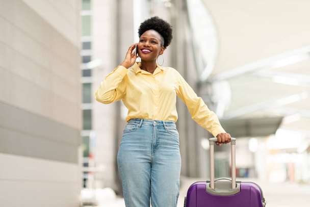 Vrolijke Afro-Amerikaanse vrouw reiziger praten over de telefoon staande met koffer in de luchthaven Terminal Binnenshuis. Reis- en mobiele communicatie bieden concept - Foto, afbeelding