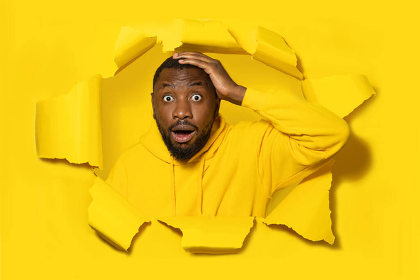 Homem negro chocado tocando sua cabeça e olhando através de buraco em papel amarelo, espaço livre. Expressão facial incrível, emoções, publicidade, venda e oferta - Foto, Imagem
