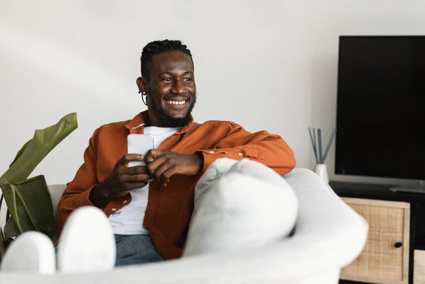 Heyecanlı siyahi adam cep telefonuyla sohbet ediyor ya da internette sörf yapıyor, evdeki koltukta oturuyor, fotokopi odasına bakıyor ve gülümsüyor. Modern teknolojiler ve yaşam tarzı - Fotoğraf, Görsel