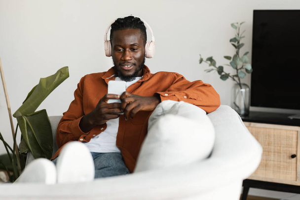Odprężony czarny człowiek oglądający filmy na smartfonie, noszący słuchawki i korzystający z telefonu komórkowego, siedzący na kanapie, kopiujący przestrzeń. Afrykański Amerykanin surfuje po Internecie i słucha muzyki w domu - Zdjęcie, obraz