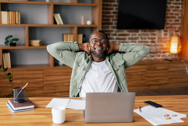 Happy successful middle aged african american man in glasses and casual in wireless headphones resting from work with laptop in home office interior. New normal and business, technology, take break - Zdjęcie, obraz