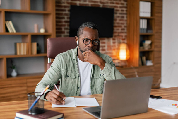 Serious concentrated adult african american man in glasses and casual in wireless headphones works on laptop and thinks over business plan in home office interior. Brainstorm, meeting and video call - Photo, Image