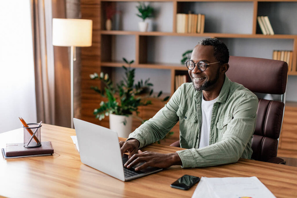 Vrolijk vertrouwen volwassen Afrikaanse Amerikaanse zakenman in bril en casual werken op laptop in huis kantoor interieur. Nieuw normaal met technologie, succesvolle zaken thuis op afstand tijdens covid-19 - Foto, afbeelding