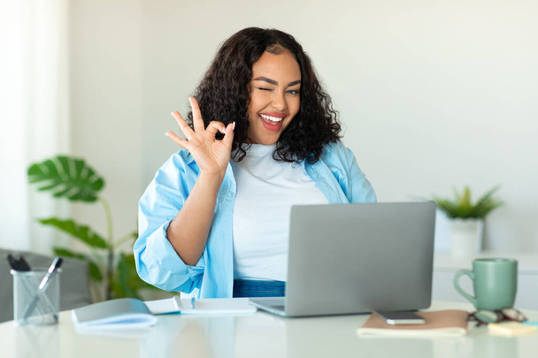 Веселый черный бизнес-женщина с помощью ноутбука Жесты Одобрение веб-сайт Поза улыбка на камеру, сидя в современном офисе. Женщина показывает хорошо знак работы в Интернете на рабочем месте в помещении - Фото, изображение