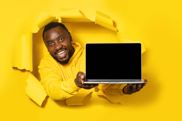 広告の無料場所。黄色い紙の引き裂かれた穴を通して装置を示す空白の画面でラップトップを保持している陽気な黒人男性。モックアップのためのコピースペースを持つガジェット,ウェブサイトテンプレート - 写真・画像