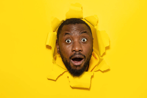 Choqué homme afro-américain réagissant avec surprise, regardant la caméra avec une expression impressionnée, posant dans un trou de papier déchiré, fond jaune - Photo, image