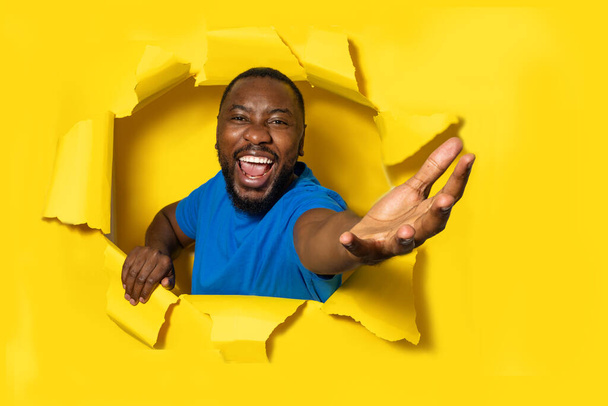Excited african american man stretching open palm through hole in torn yellow paper, taking or giving something, holding invisible object - Photo, Image