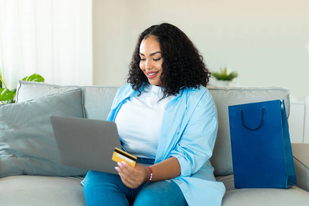 Μαύρη γυναίκα αγοραστής ψώνια σε απευθείας σύνδεση χρησιμοποιώντας το laptop και την πιστωτική κάρτα αγορά συν μέγεθος ρούχα στην αγορά Web συνεδρίαση στο σπίτι. Internet Banking και Ecommerce Concept - Φωτογραφία, εικόνα