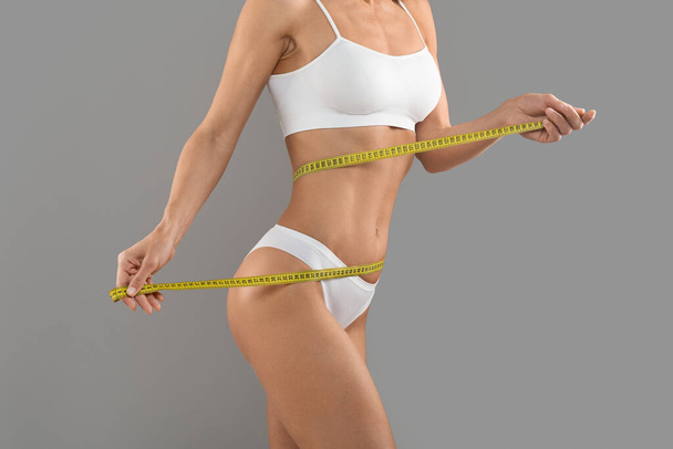 Healthy lifestyle concept. Fit woman in underwear measuring her waistline with measure tape while posing isolated over grey background, unrecognizable young female with sporty body checking measures - Photo, Image