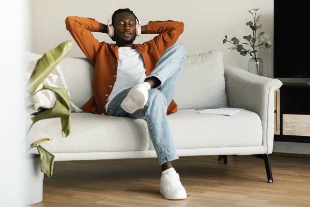 Descanso fin de semana. Hombre negro relajado en auriculares inalámbricos escuchando audiolibro en casa, sentado con los ojos cerrados en el sofá en la sala de estar, disfrutando del ocio doméstico, espacio para copiar - Foto, imagen