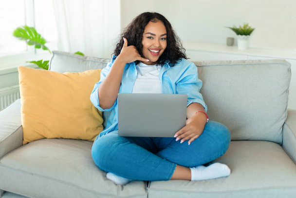 ラップトップを使用して幸せなアフリカ系アメリカ人女性とジェスチャーカメラの仕事に笑顔を呼び出し、自宅でソファに座ってオンライン通信.フリーランスと距離通信 - 写真・画像
