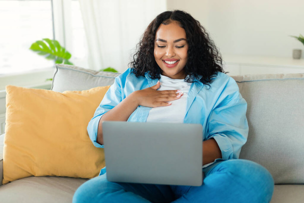 Cheerful Black Lady Using Laptop Computer Watching Movie Sitting On Sofa At Home. Freelancer Woman Working, Communicating And Learning Online. Internet Technology And Freelance Concept - 写真・画像