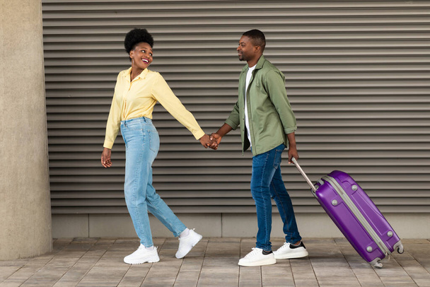 Ταξίδια διακοπών. Ευτυχισμένο ζευγάρι Αφροαμερικανών τουριστών που περπατούν με βαλίτσα κρατώντας τα χέρια έξω κοντά στον τερματικό σταθμό του αεροδρομίου. Τουρισμός και ταξίδια Concept. Πλήρες μήκος, πλευρική άποψη shot - Φωτογραφία, εικόνα