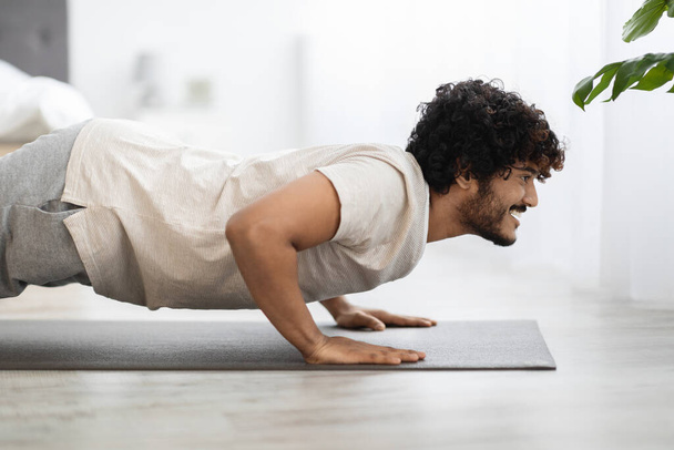 Positieve bebaarde jonge oosterse man in sportkleding die thuis op fitnessmat traint, indiaanse man die 's ochtends yoga oefent, kijkend naar kopieerruimte voor reclame, slaapkamerinterieur, zijaanzicht - Foto, afbeelding