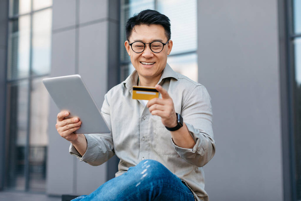 Tecnologia, pagamento online e conceito de pessoas. Empresário coreano feliz com tablet digital e cartão de crédito encomendar bens ao ar livre, sentado perto do prédio de escritórios - Foto, Imagem