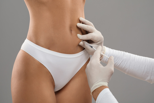 Beautician Doctor Wearing Gloves Making Beauty Injection To Female Patient Belly, Unrecognizable Young Woman In Underwear Getting Lipolysis Treatment For Slimming, Standing On Grey Background - Foto, immagini