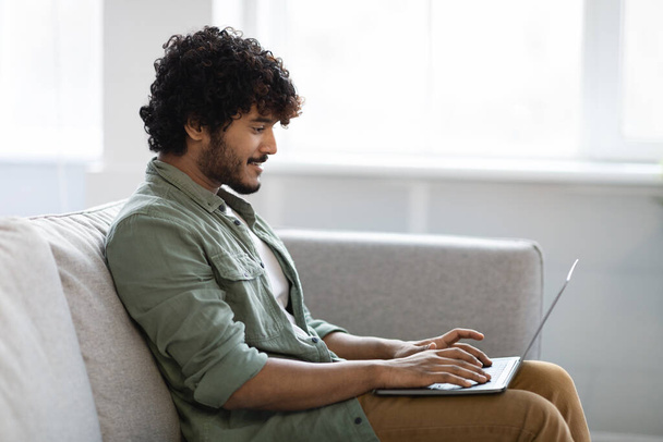 Side view of positive indian guy in casual outfit sitting on couch at home, using modern laptop, typing on computer keyboard and smiling, freelancer enjoying his remote job, copy space - Photo, Image