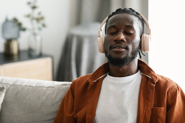 Kablosuz kulaklıklı sakin Afro-Amerikan adam kanepeye oturmuş, gözleri kapalı evde müzik dinliyor, boş yer arıyor. Zenci adam kulaklıkla yeni ses kitabının keyfini çıkarıyor. - Fotoğraf, Görsel