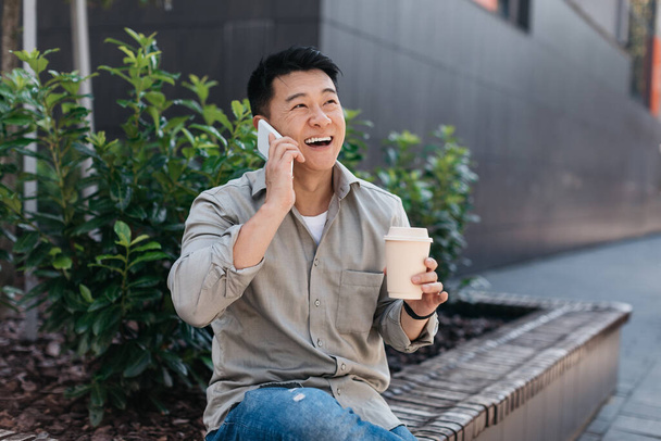 Happy asian male entrepreneur talking on smartphone and drinking coffee from takeaway cup, sitting outdoors near modern office building. Businessman using gadget for remote business - Photo, Image