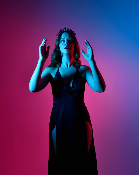 Portrait en gros plan d'une belle mannequin femme portant une élégante robe noire et une couronne, posant sur un fond de studio avec des gestes de bras inspirés de fantaisie, un éclairage créatif multicolore. - Photo, image