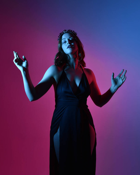 Close up portrait of beautiful woman model wearing elegant black dress and crown, posing against a studio background with fantasy inspired arm gestures, multi coloured creative lighting. - Photo, image