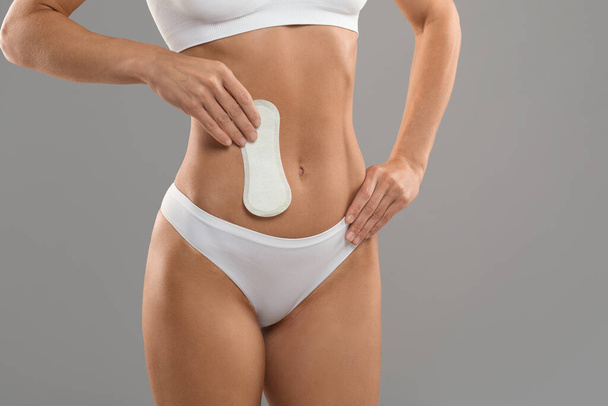 Female Hygiene. Young unrecognizable lady in white underwear holding clean menstrual pad in hand near abdomen while standing isolated over grey studio background, cropped, copy space - Foto, afbeelding