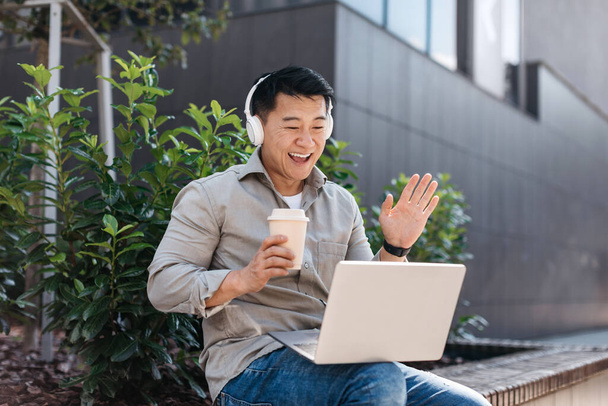 Vzrušený asijský podnikatel středního věku pracující venku na notebooku, ve sluchátkách a s videohovory, pije kávu na lavičce poblíž kancelářské budovy. Obchod venku, přestávka na kávu - Fotografie, Obrázek