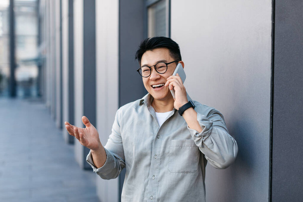 Empresario masculino asiático que llama por teléfono inteligente y gesto, de pie cerca del moderno edificio de oficinas, al aire libre. Jefe conversando con el cliente por negocios - Foto, imagen
