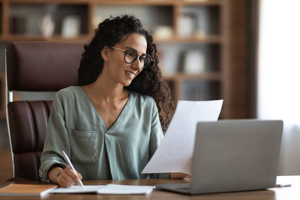 Hübsche junge brünette Geschäftsfrau sitzt am Schreibtisch vor dem Laptop, überprüft Korrespondenz, liest Dokumente, trägt eine Brille, arbeitet im Büro, Kopierraum - Foto, Bild