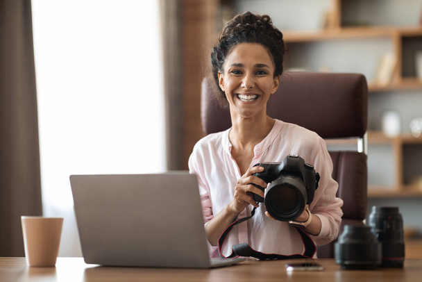 Позитивний красивий фотограф, що працює вдома, сидить на робочому столі з сучасним ноутбуком, тримає професійну камеру, п'є каву, перевіряє фотографії, копіює простір
 - Фото, зображення