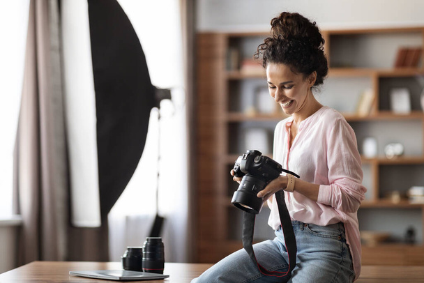 Veselá atraktivní mladá dáma v neformálním oblečení fotograf sedí na stole ve studiu nebo kanceláři, drží profesionální fotoaparát, kontrola fotografií a s úsměvem, kopírovací prostor. Koncept pracovních míst a povolání - Fotografie, Obrázek