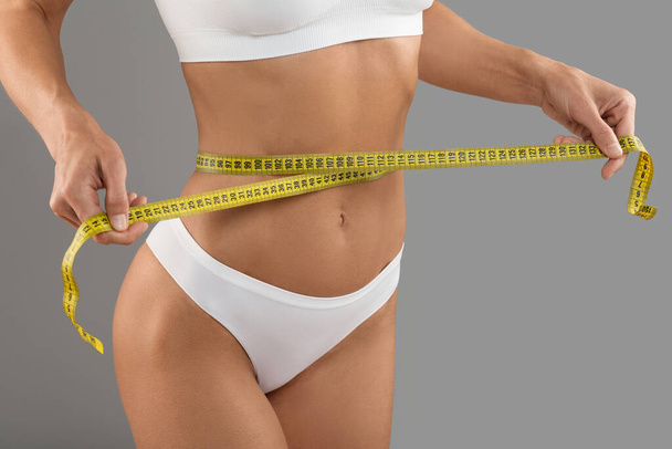 Nahaufnahme einer jungen Frau mit Maßband zur Überprüfung der Taillengröße nach der Diät, unkenntlich schlanke Frau mit fittem Körper in weißer Unterwäsche, die über grauem Studiohintergrund posiert, beschnitten - Foto, Bild