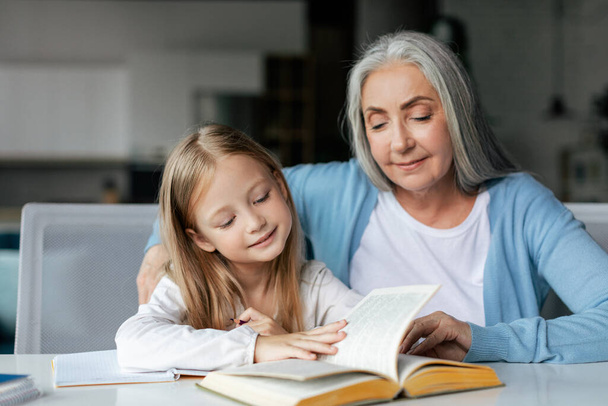 Smiling concentrated smart european small girl with retired grandmother read book, enjoy study and spare time in living room interior. Relationship and care, education and lesson at home together - Fotó, kép