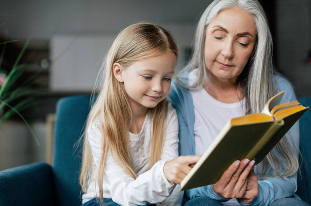 Serious caucasian small granddaughter and mature grandmother are reading book on sofa in room interior, close up. Love and family relationship, study together, bedtime story and education at home - Fotó, kép