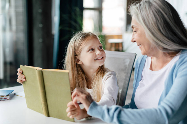 Cheerful caucasian little preschooler with aged grandma read book, enjoy study and free time in living room interior. Education and lesson together at home, new normal during covid-19 quarantine - Foto, afbeelding