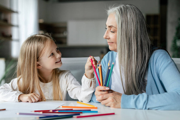 Lächelnde europäische Enkelin mit alter Großmutter mit bunten Bleistiften malen ein Bild auf den Tisch, genießen Studium und Freizeit im Wohnzimmerinterieur. Bildung und Kunstunterricht gemeinsam zu Hause - Foto, Bild