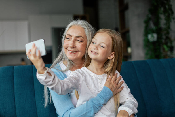 Happy caucasian little granddaughter and elderly grandma make selfie on phone, have video call on sofa in living room interior. Family relationship, lifestyle and communication with gadget at home - Photo, Image