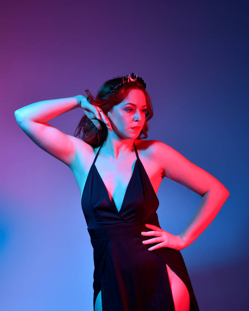 Close up portrait of beautiful woman model wearing elegant black dress and crown, posing against a studio background with fantasy inspired arm gestures, multi coloured creative lighting. - Fotó, kép