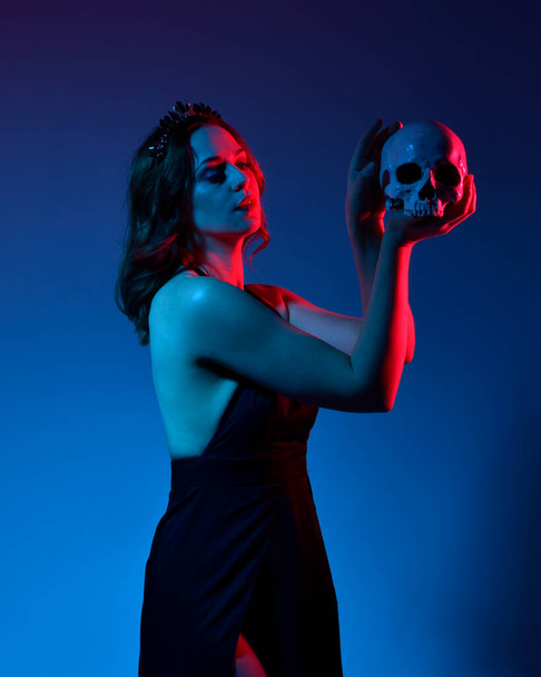 Close up portrait of beautiful woman model wearing elegant black dress and crown, posing against a studio background with fantasy inspired arm gestures holding a human skull prop.  multi coloured creative lighting. - Фото, зображення