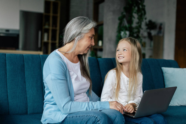 Smiling caucasian small granddaughter and old grandma have video call on computer, study online, play on sofa in living room interior. Love and family relationship, new normal due covid-19 quarantine - Foto, afbeelding
