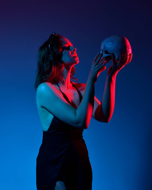 Close up portrait of beautiful woman model wearing elegant black dress and crown, posing against a studio background with fantasy inspired arm gestures holding a human skull prop.  multi coloured creative lighting. - Foto, Imagen