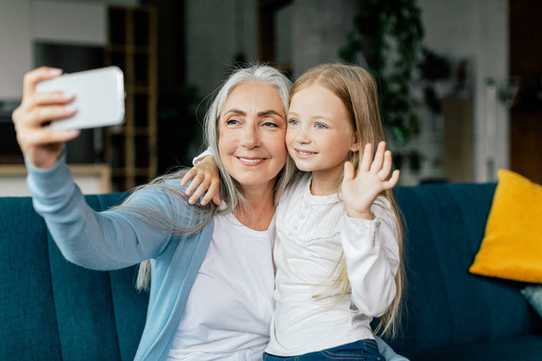 Glad caucasian small girl and retired granny waving hand in smartphone, have video call on sofa in living room interior. Remote communication, gadget for blog, new app during covid-19 quarantine - Foto, Imagen