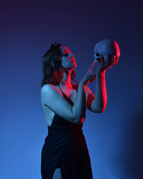 Close up portrait of beautiful woman model wearing elegant black dress and crown, posing against a studio background with fantasy inspired arm gestures holding a human skull prop.  multi coloured creative lighting. - Foto, Bild