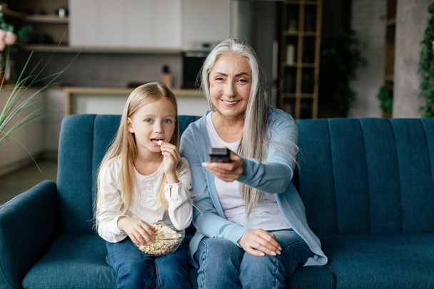 Happy caucasian small girl eat popcorn with elderly lady with remote control, watch tv on sofa in living room interior. Relax, entertainment in spare time, movie evening, cartoons together at home - Photo, Image