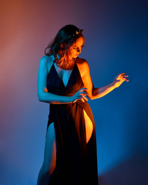 Retrato de cerca de la hermosa modelo de mujer con elegante vestido negro, posando sobre un fondo de estudio con gestos de brazos inspirados en la fantasía, iluminación creativa multicolor. - Foto, imagen